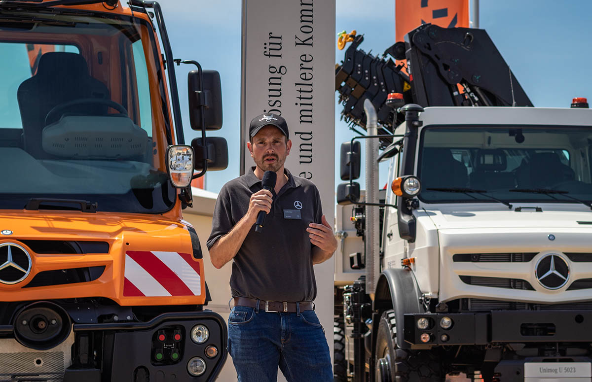 Steffen Voelckel mit Mikrofon spricht vor zwei geparkten Fahrzeugen, genauer Unimog Geräteträger