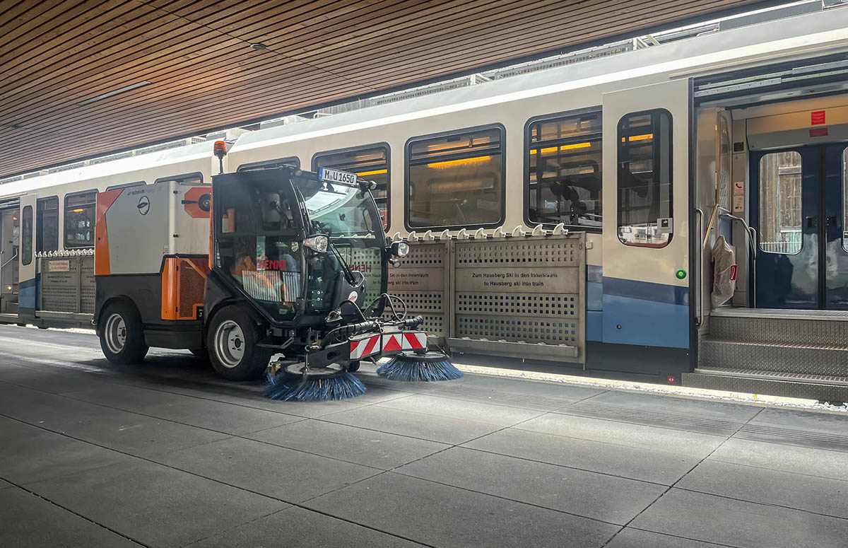 Die Kehrmaschine Flexigo150 steht auf dem Bahnsteig der bayerischen Zugspitzbahn, dahinter die Bahn selbst mit geöffneten Türen.